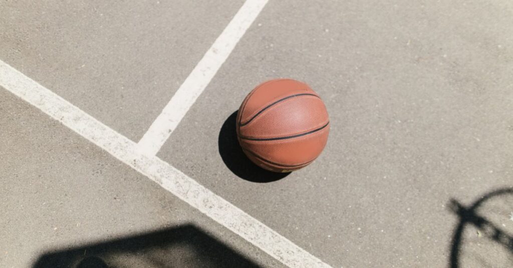 A lone basketball on a sunlit outdoor court with a hoop shadow.